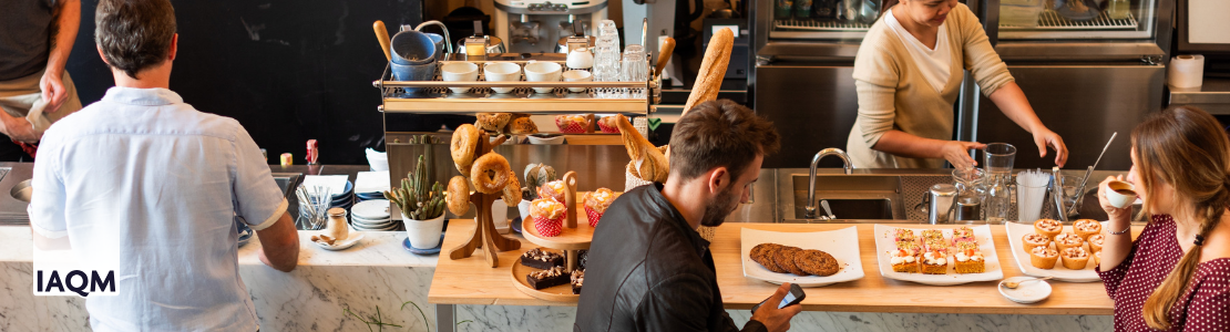 A busy cafe with people sat at atbles enjoying coffee and pastries, a man placing an order at the counter, and staff working to prepare oders.